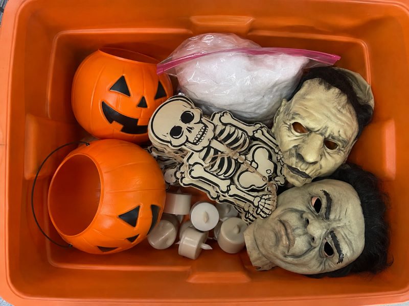Top-down photo of an open orange Halloween tote: two plastic pumpkin pails, a cardboard skeleton garland, a bag of spider webbing, two latex masks and a pile of LED tea-light candles