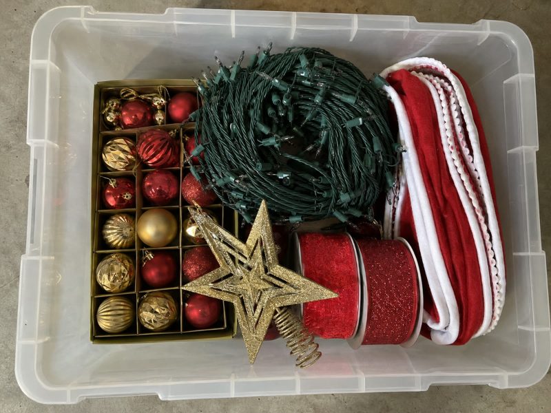 Top-down photo of an open holiday tote: glass ornaments in a divided tray, a coiled string of lights, red garland and a gold tree-topper star